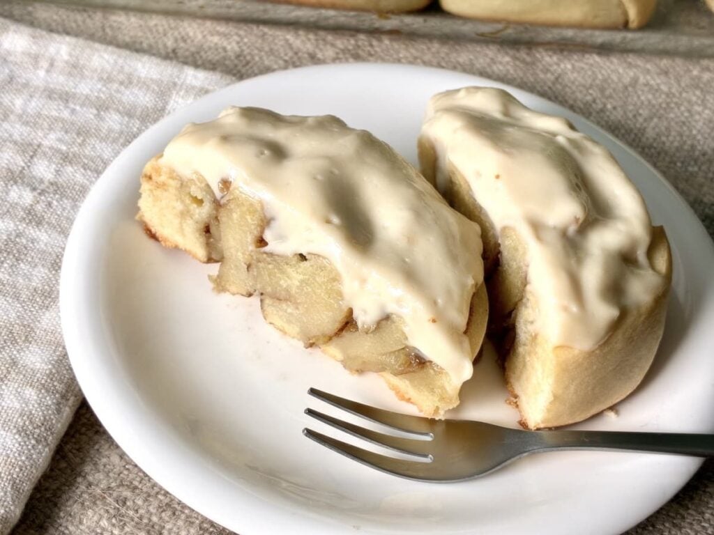 Two cinnamon rolls with creamy icing on a white plate, next to a fork and a textured beige napkin.