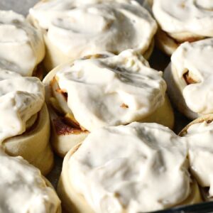 A close-up of several freshly baked cinnamon rolls topped with creamy white icing, closely packed together in a baking dish.