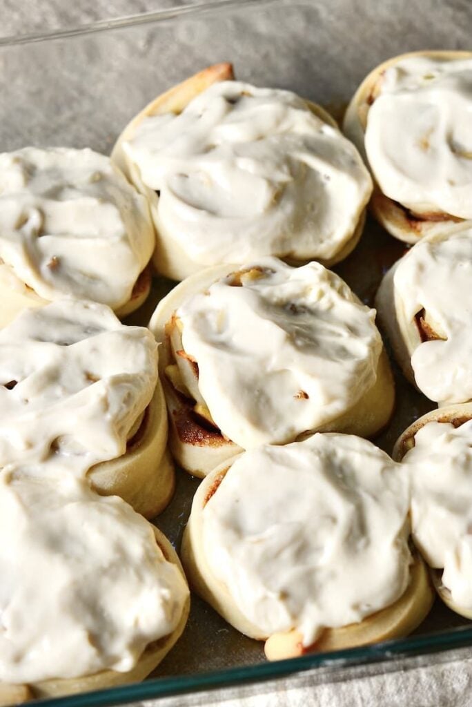 Nine cinnamon rolls topped with creamy white icing are arranged closely together in a glass baking dish. The rolls are soft and swirled, showing some cinnamon filling peeking through the icing.