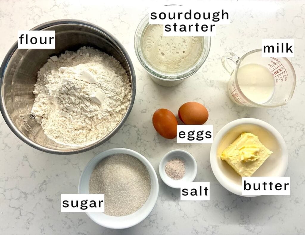 A bowl of flour, a jar of sourdough starter, a measuring cup of milk, two eggs, a dish of sugar, a dish of salt, and a dish of butter on a white counter. Each ingredient is labeled.