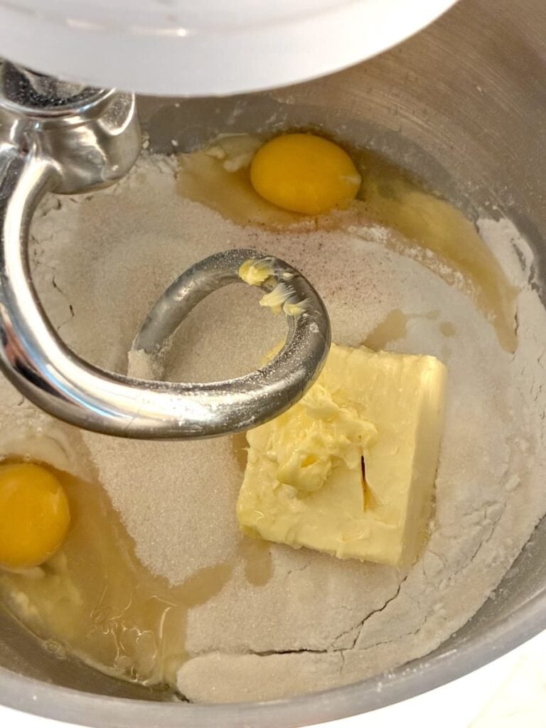 Stand mixer bowl with flour, sugar, two eggs, and a block of butter, ready to be mixed with a dough hook attachment.
