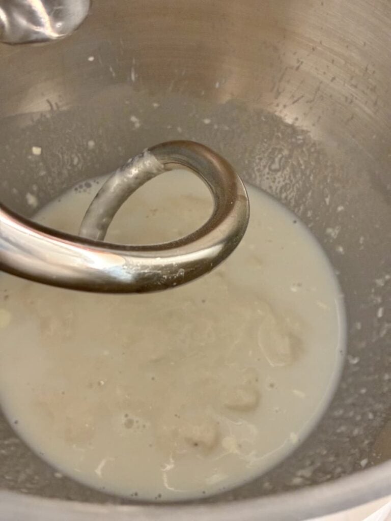 A metal dough hook in a mixing bowl with a milky liquid and lumps, possibly yeast and flour, beginning to mix for dough preparation.
