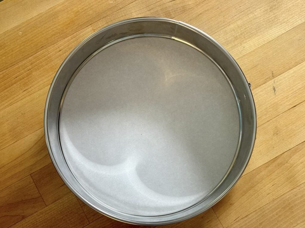 A round metal cake pan lined with a neatly cut circle of white parchment paper sits on a wooden surface.