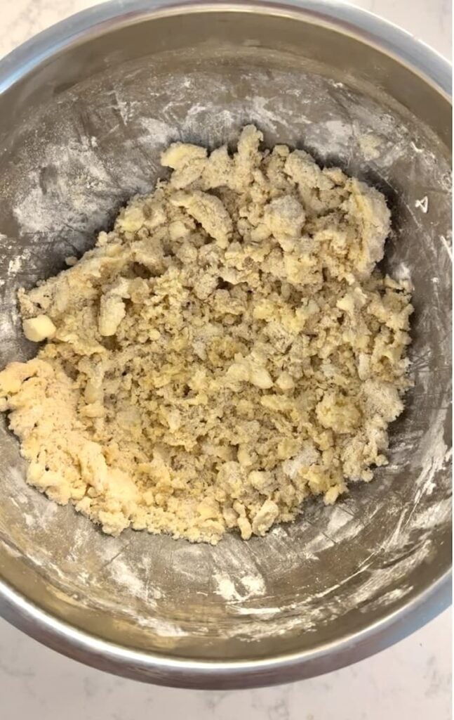 A metal bowl filled with coarse, crumbly dough mixture for apple crumb cake sits on a light countertop. Some flour is visible on the sides of the bowl.