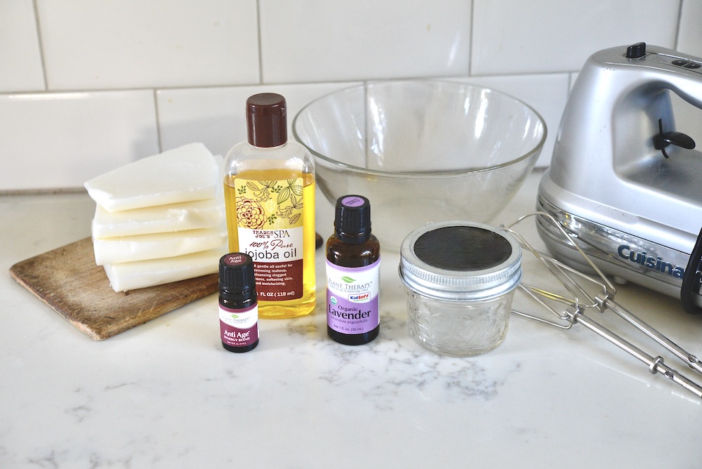 beef tallow, jojoba oil, glass bowl, hand mixer, whisk attachments, glass jar, lavender essential oil, and anti-age essential oil on kitchen counter