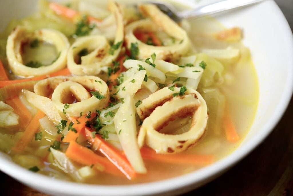 A bowl of vegetable soup with sliced carrots, cabbage, herbs, and round, grilled pieces resembling those in a German pancake soup, all in a clear broth with a spoon resting on the side.