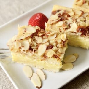 Three squares of almond-topped pastry filled with creamy custard are served on a white plate with sliced almonds and two strawberries, with a fork resting on the side.