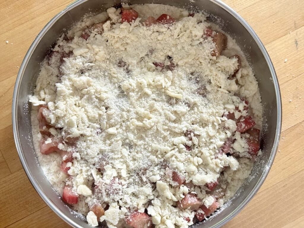 A round metal pan filled with chopped rhubarb pieces topped with an uncooked, crumbly streusel topping sits on a wooden surface, ready to be baked.