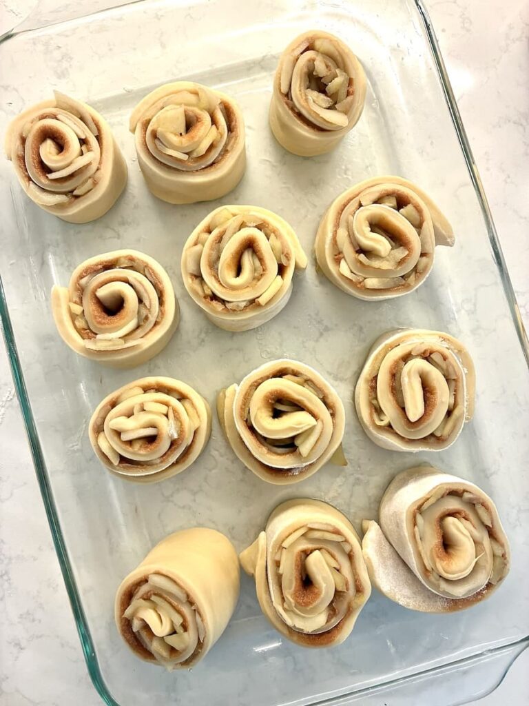 Unbaked cinnamon rolls arranged in a glass baking dish on a marble countertop, showing their spiral layers of dough and cinnamon filling.