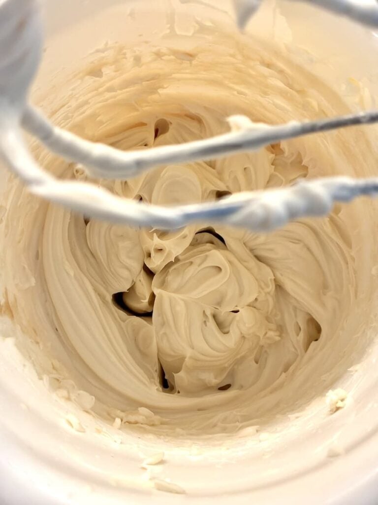 A close-up of creamy, whipped frosting inside a mixing bowl, with two beaters visible at the top coated in the frosting.