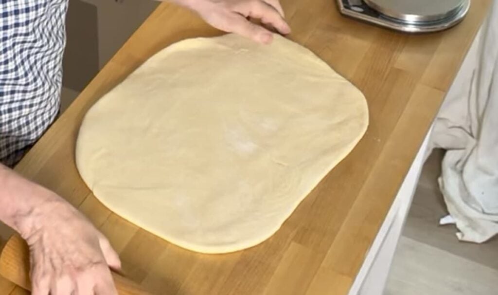 A person rolls out a sheet of dough on a wooden countertop using a rolling pin, preparing to make German butterkuchen, with a digital kitchen scale visible in the background.
