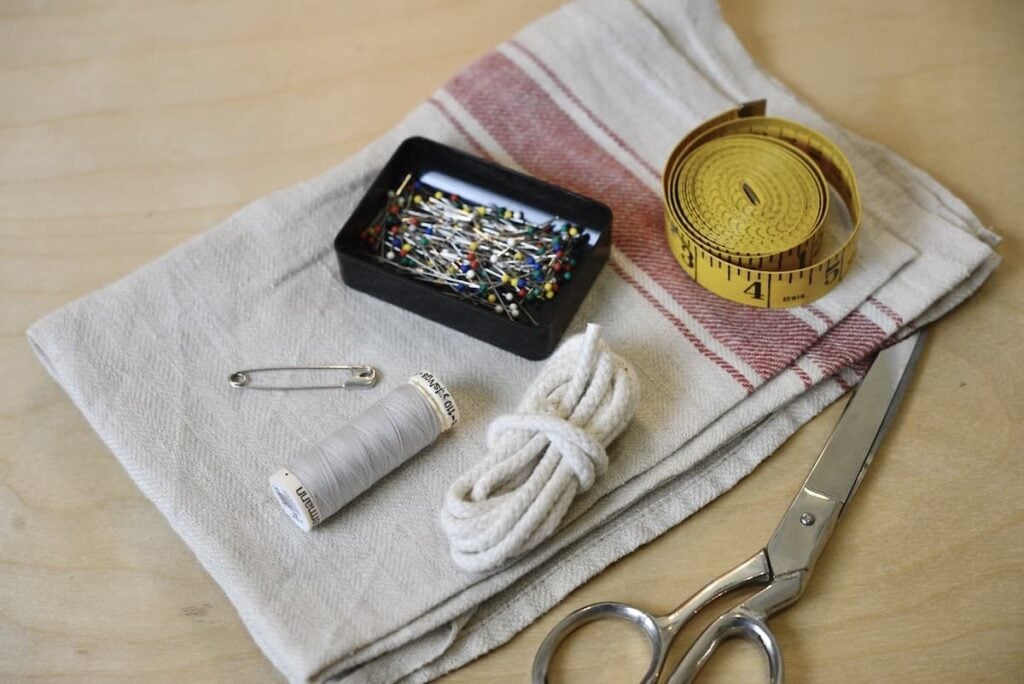 A folded linen bread bag, a spool of white thread, straight pins in a container, a safety pin, a coiled measuring tape, a bundle of white cord, and metal scissors rest on a wooden surface.