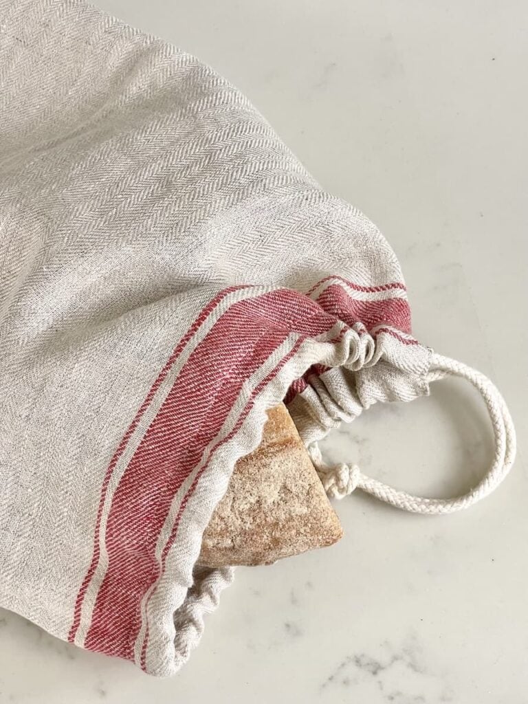 A loaf of bread partially wrapped in a linen bread bag with red stripes and a drawstring, resting on a light-colored, speckled surface.