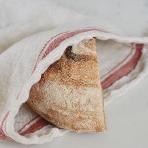 A rustic loaf of bread, partially wrapped in a linen bread bag with red stripes, rests on a light-colored surface.
