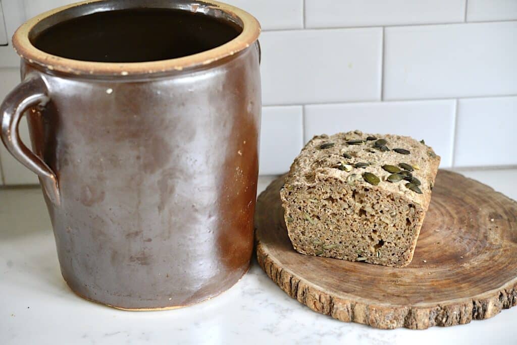 bread on a wooden cutting board next to a stone crock on kitchen counter