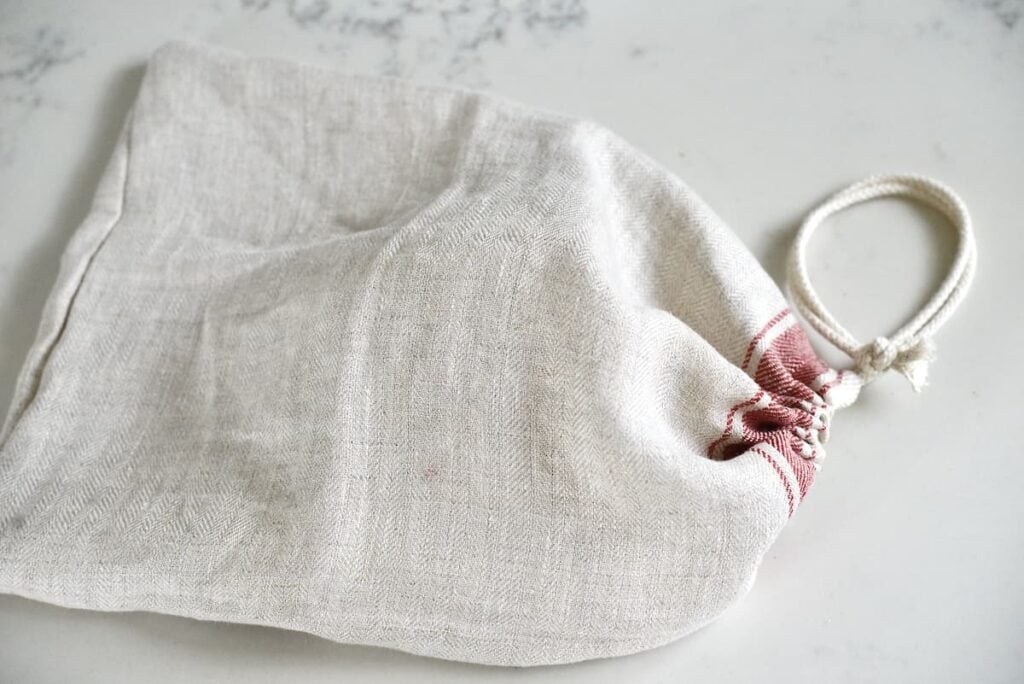 A beige linen bread bag with red stitching and a white cord, resting on a light-colored surface.