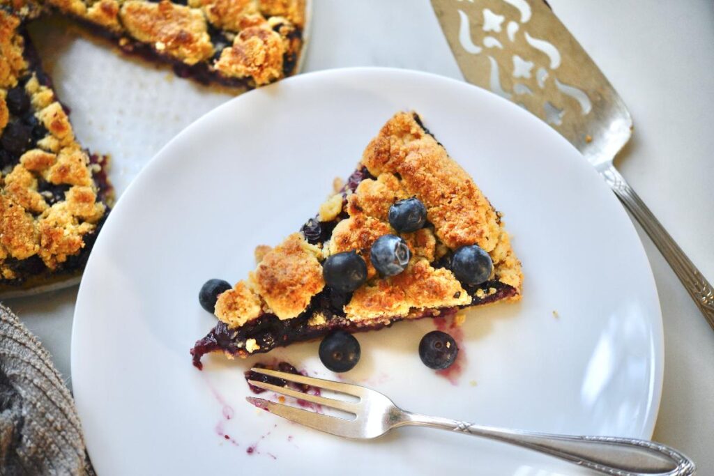 a slice of German blueberry Streuselkuchen on a white plate with fork, next to the rest of the cake and a pie server