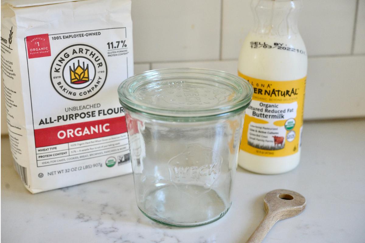 A package of King Arthur Flour organic, unbleached flour, a Weck glass jar, container of buttermilk, and a wooden spoon on kitchen counter.