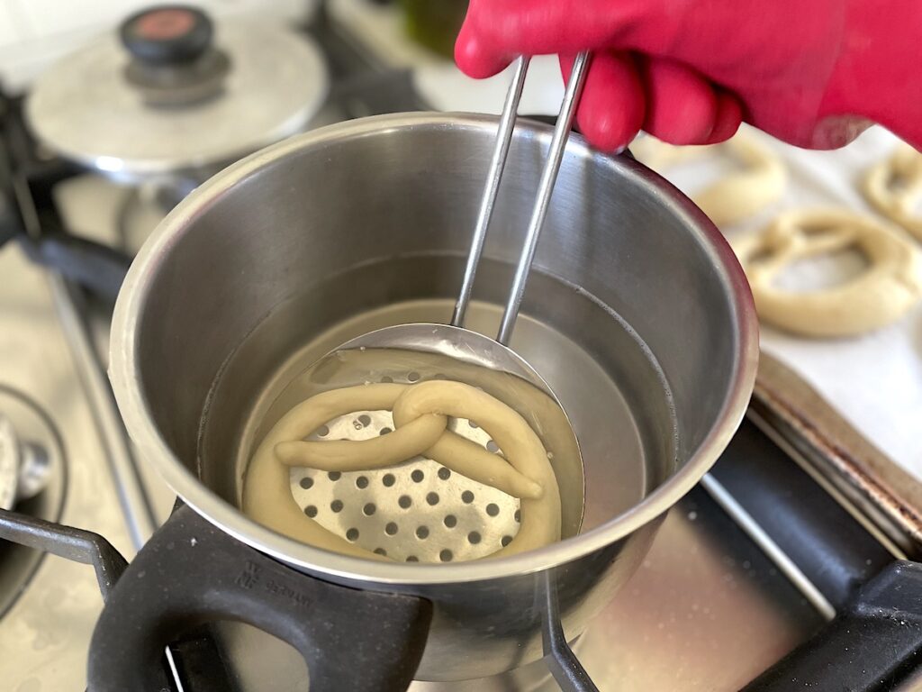 unbaked pretzel on slotted spoon in pot with lye solution