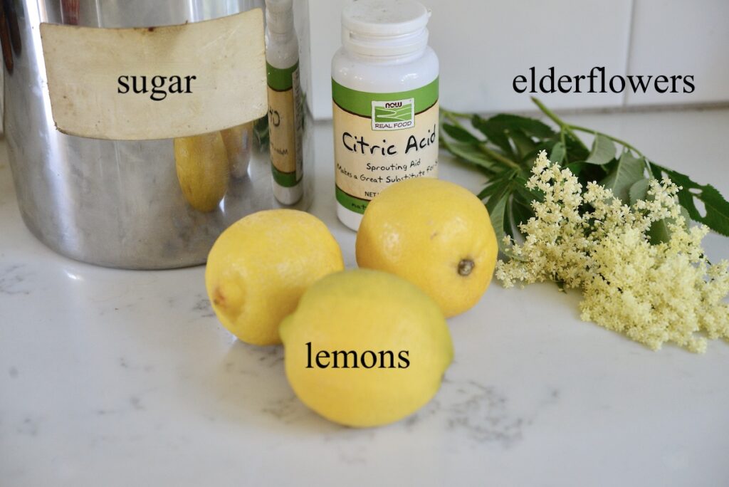jar of sugar, bottle of citric acid, elderflowers, and 3 lemons on kitchen counter