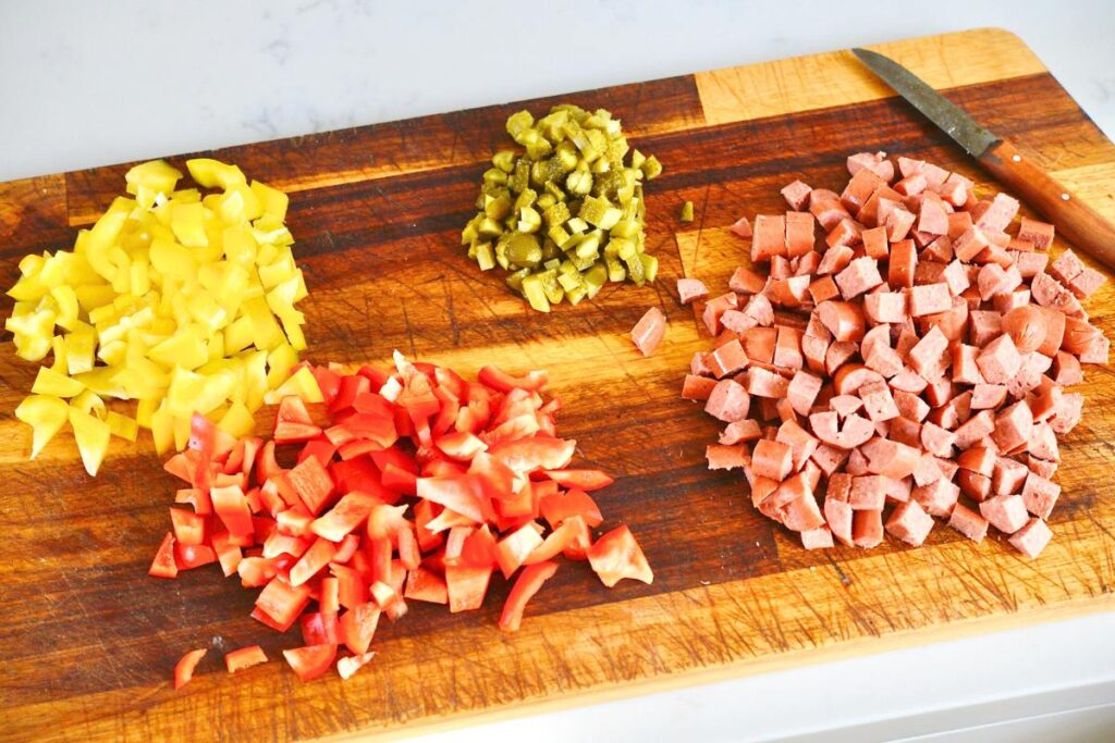 wooden chopping board with chopped yellow bell peppers, red bell peppers, cornichons, and hot dogs
