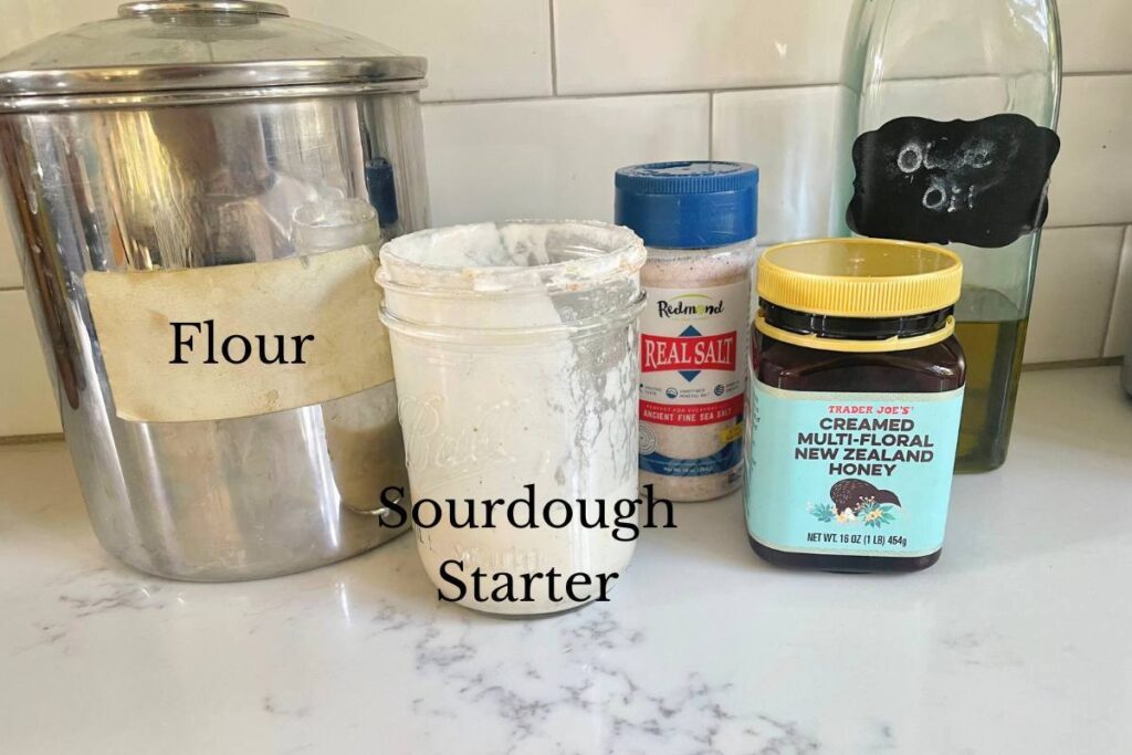 flour, jar of sourdough starter, salt, honey, and olive oil on kitchen counter