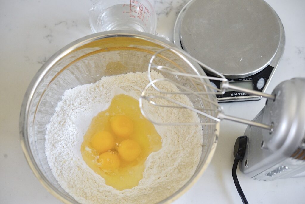flour and eggs in mixing bowl with electric hand mixer and kitchen scale