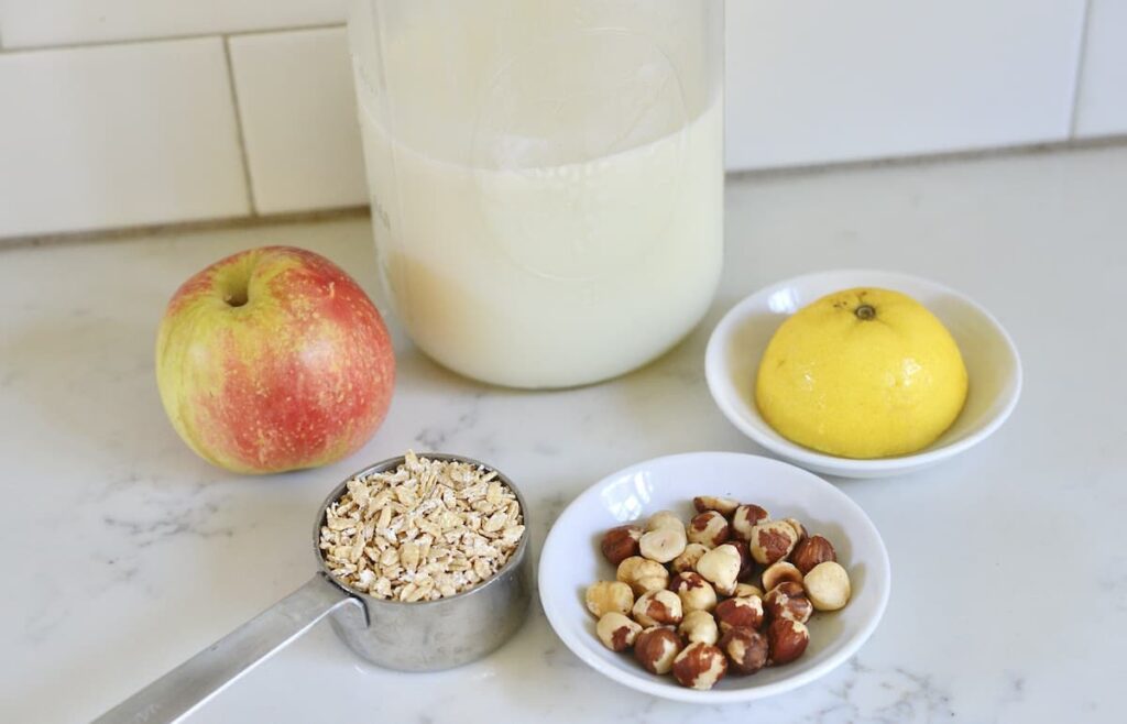 bottle of milk, an apple, rolled oats, whole hazelnuts, and half a lemon on kitchen counter