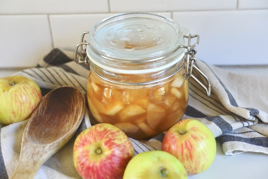 canning jar with apple pie filling on kitchen counter with apples, wooden spoon, and tea towel