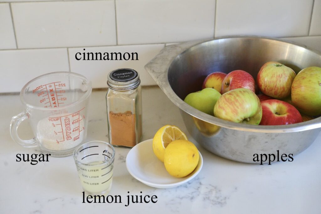 measuring cup with sugar, jar of ground cinnamon, cup with lemon juice and halved lemon, and bowl with apples on kitchen counter