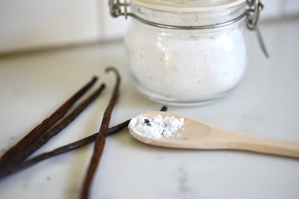 homemade vanilla sugar on a wooden spoon in front of jar and with vanilla beans