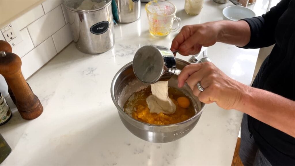Woman's hands adding a measuring cup of wet ingredients to a mixing bowl of dry ingredients.