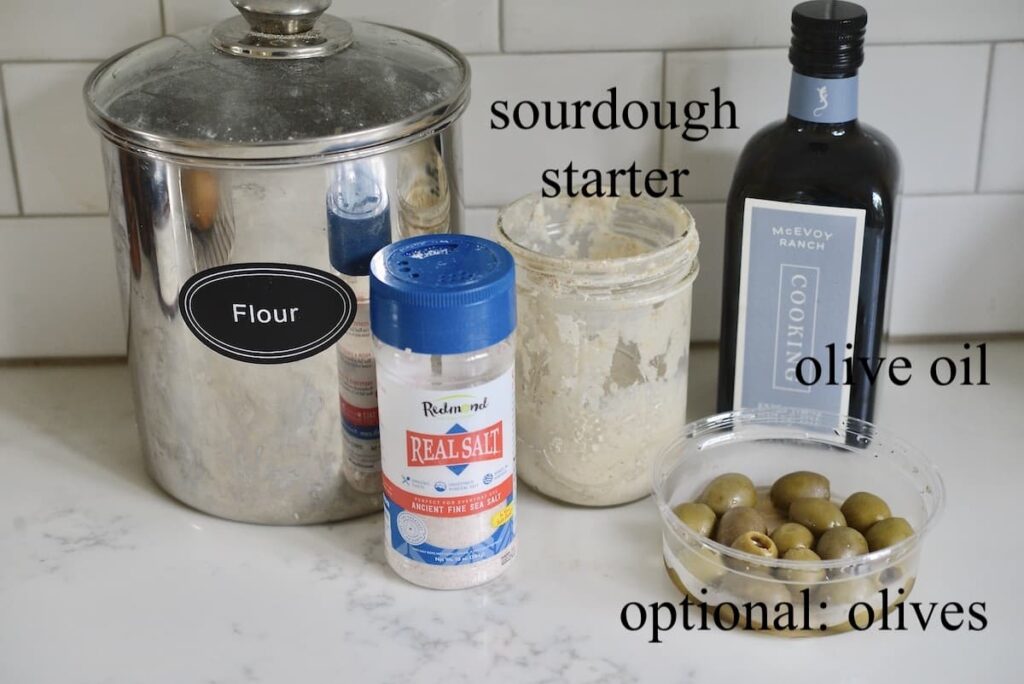 flour, sea salt, jar of sourdough starter, olive oil, and green olives on kitchen counter