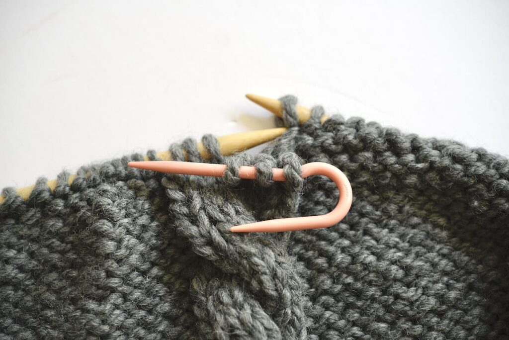 knitted piece with knitting needles showing how to pull the back stiches forward