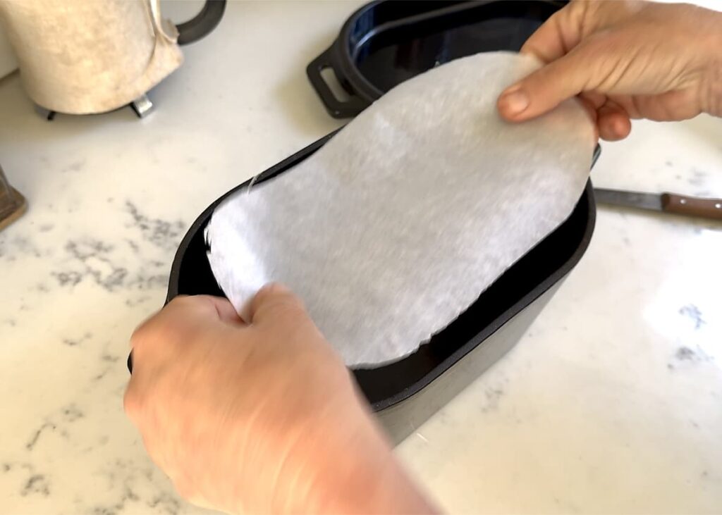 hands placing oval piece of parchment paper in oval Dutch oven