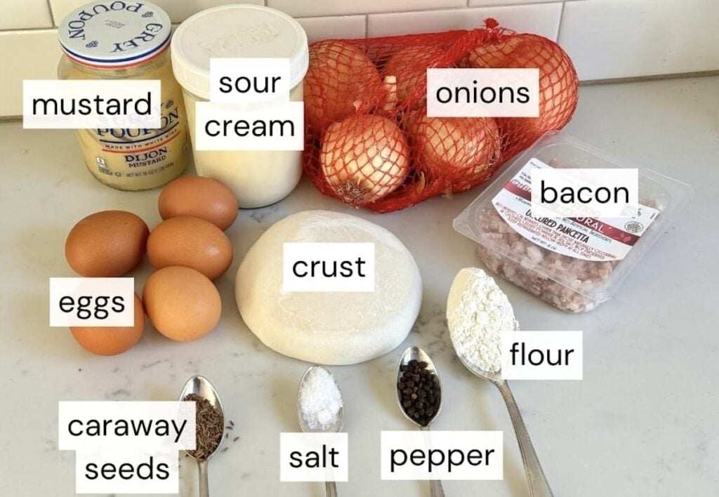 German onion pie ingredients on kitchen counter: mustard, sour cream, onions, bacon, crust, eggs, caraway seeds, salt, black pepper, and flour
