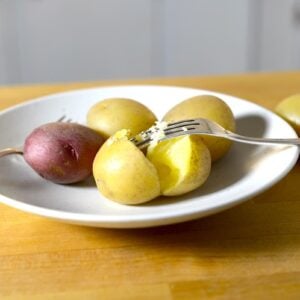 A white plate with several small boiled potatoes, both red and yellow varieties, on a wooden surface. Wondering how long to boil potatoes? One yellow potato is split open with a fork beside another fork resting on the plate.
