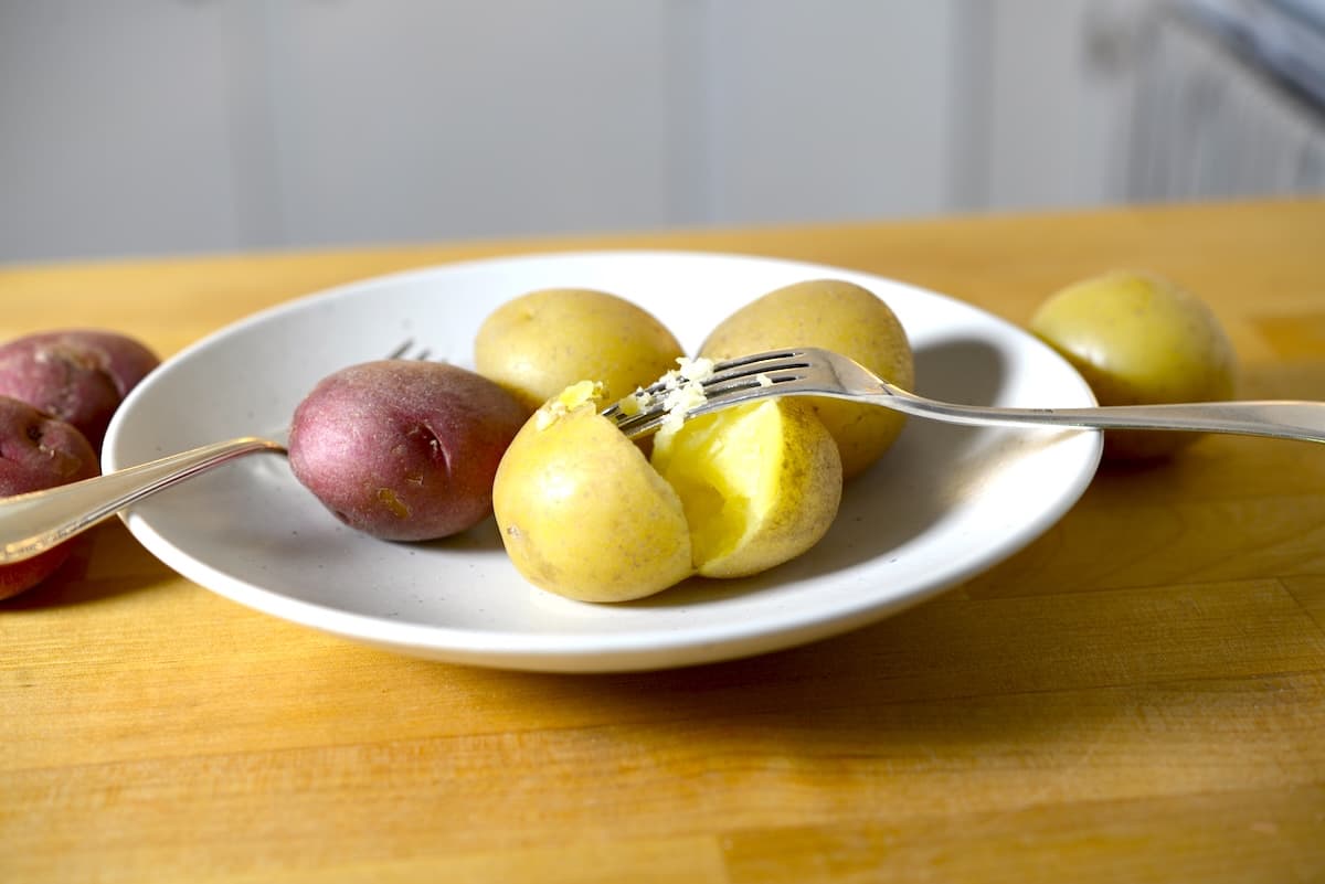 A white plate with several small boiled potatoes, both red and yellow varieties, on a wooden surface. Wondering how long to boil potatoes? One yellow potato is split open with a fork beside another fork resting on the plate.