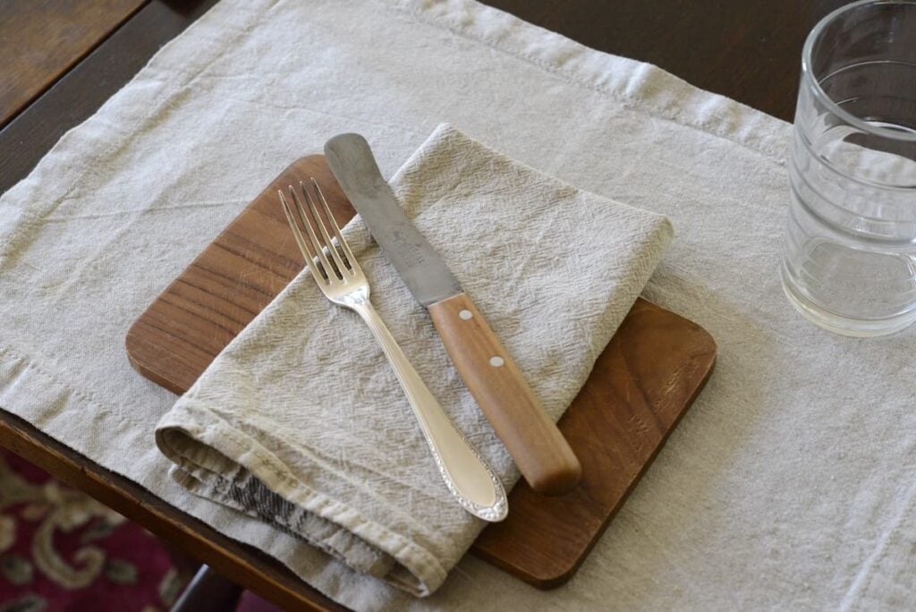 table setting with placemat, wooden board, napkin, fork, knife, and drinking glass