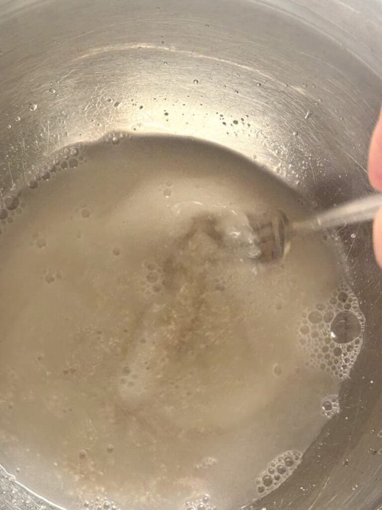 A hand holding a whisk stirs a cloudy liquid with bubbles in a metal bowl, preparing the dough for traditional German Bauernbrot.