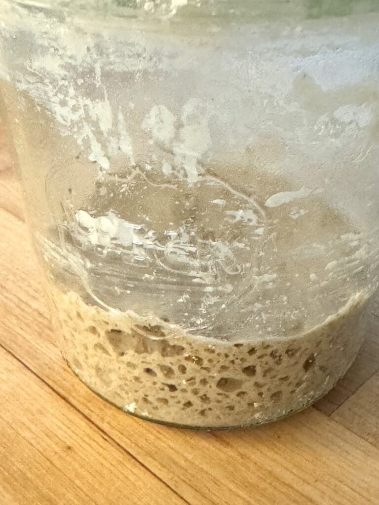 A glass jar with bubbly, active sourdough starter at the bottom sits on a wooden surface. Remnants of dried starter cling to the sides—perfect for baking a classic German Bauernbrot.