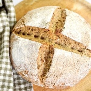 Round loaf of German Bauernbrot with a dusting of flour and a cross-shaped slash on top, placed on a wooden board next to a green and white checkered kitchen towel.