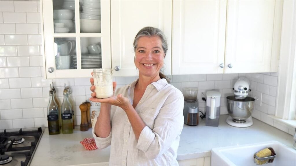 A smiling woman in a light shirt stands in a bright kitchen, holding up a glass jar with a white substance inside. Kitchen appliances, bottles, and plates are neatly arranged in the background.