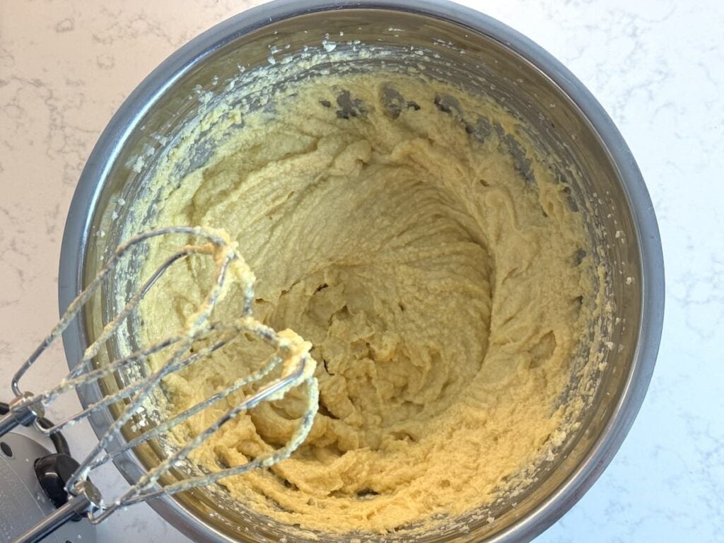A metal mixing bowl filled with pale yellow cake batter, with an electric hand mixer resting on the edge. The bowl is on a white marble countertop.