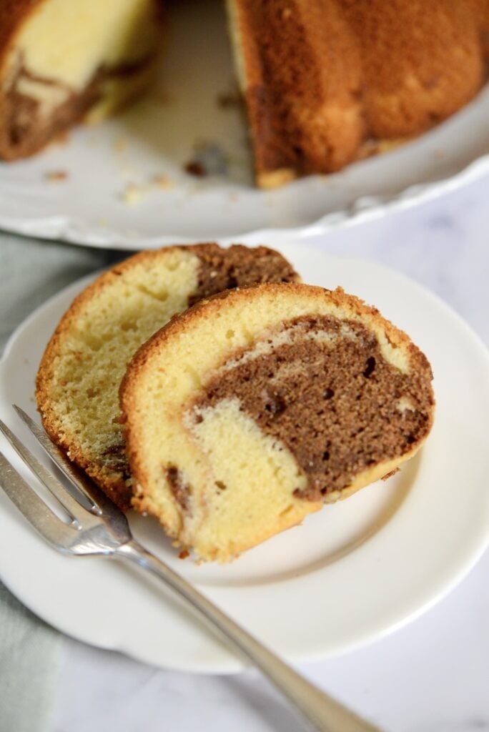 Two slices of marble cake, showing swirls of vanilla and chocolate, are served on a white plate with a fork. More cake is visible in the background.