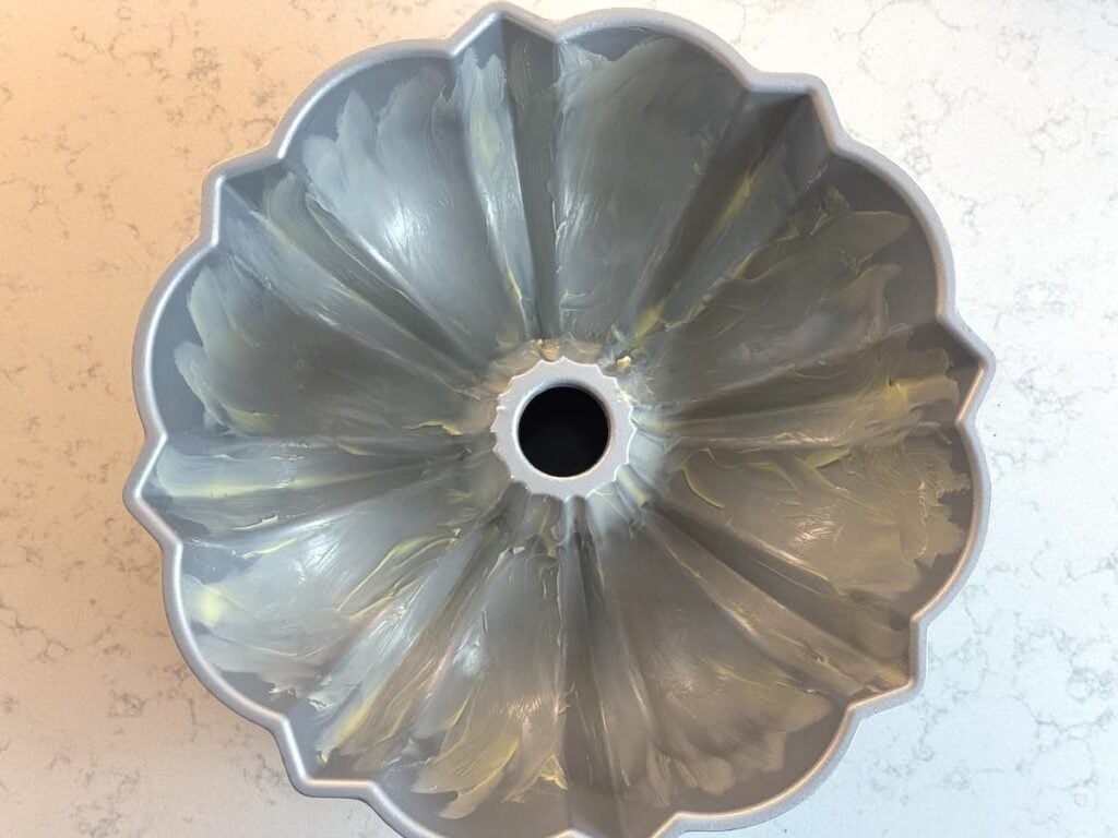 A metal Bundt cake pan with a fluted design is coated with a layer of butter, ready for baking, and sits on a light-colored marble countertop.