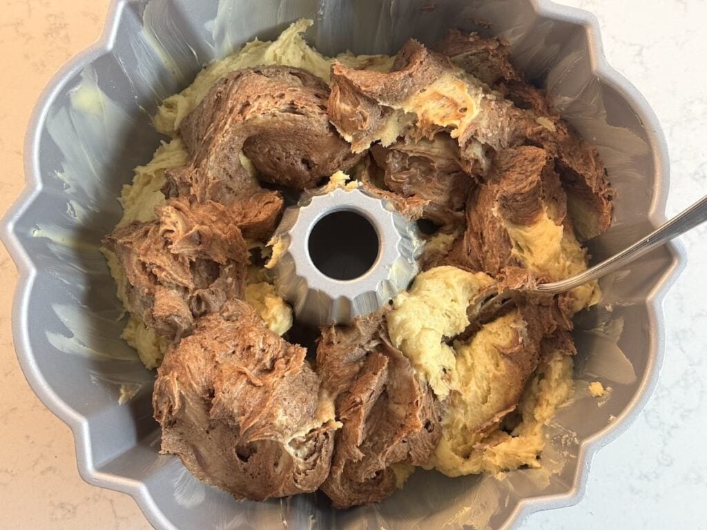 A Bundt pan filled with swirled chocolate and vanilla cake batter, ready to be baked. A fork rests in the pan, suggesting the batter is being mixed or swirled.