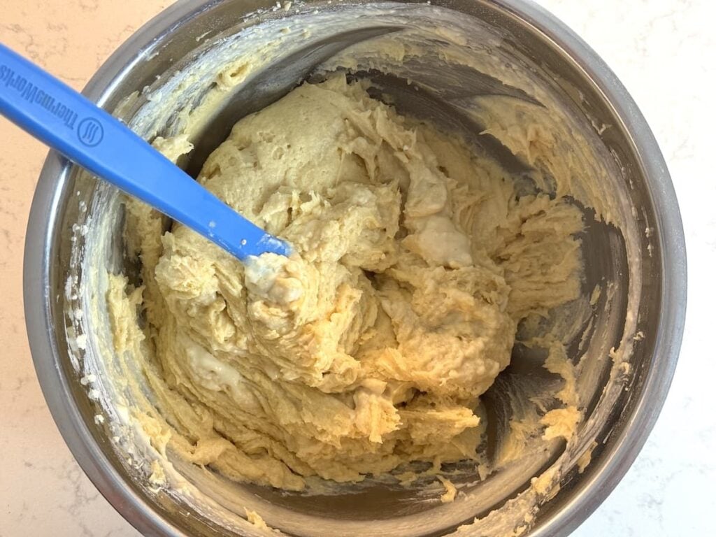 A mixing bowl filled with thick, creamy dough and a blue spatula resting inside, on a light-colored countertop.