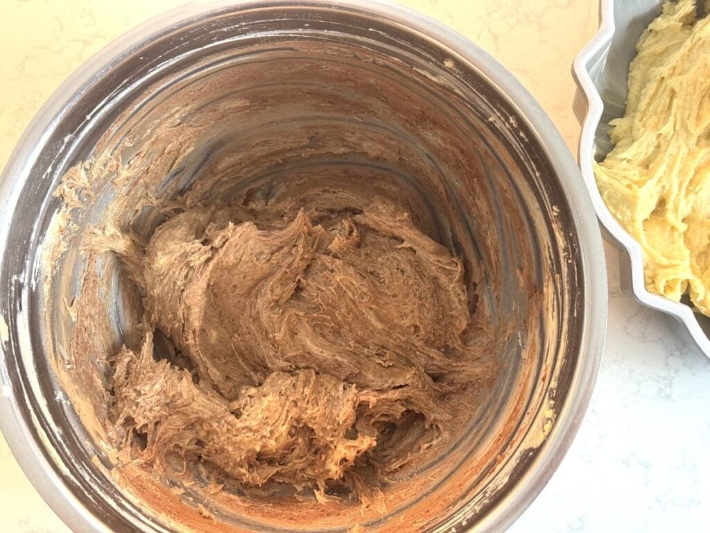 A metal mixing bowl filled with thick brown cake batter sits on a light countertop, next to a scalloped baking pan containing yellow batter.