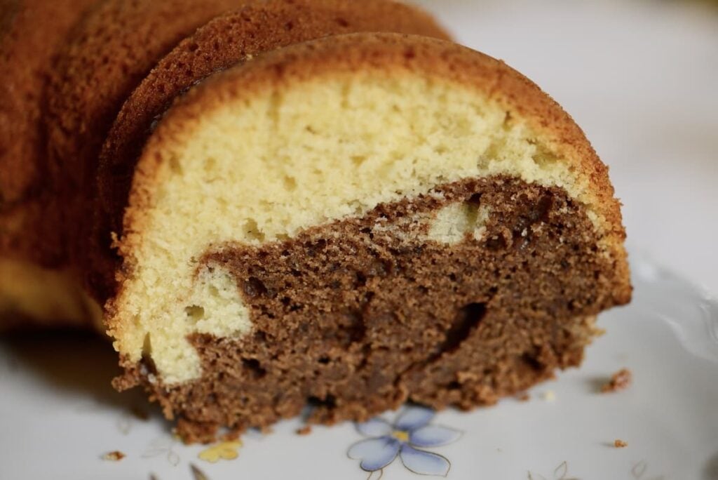 Close-up of a slice of marble cake on a white plate with a blue flower design, showing a mix of yellow vanilla and brown chocolate swirled layers.